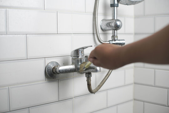 Person cleaning a dirty shower faucet with a brush in a modern bathroom with white subway tiles, emphasizing home maintenance and hygiene.