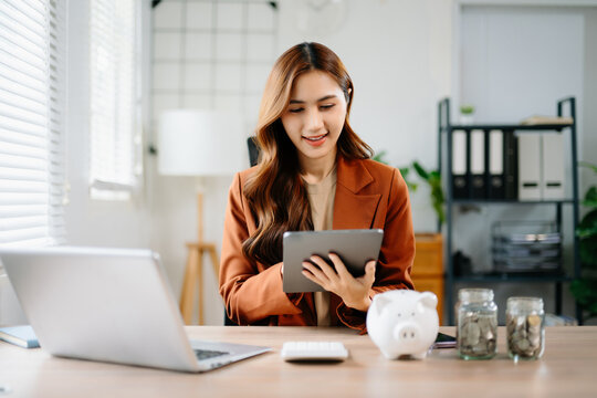 Young Asian businesswoman using tablet for finance and savings planning in modern office with piggy bank and coin jars, symbolizing money management, fintech, and success.