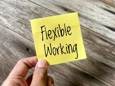 A hand holding a yellow sticky note with the phrase 'Flexible Working 'on a wooden background. Represents workplace flexibility and modern employee benefits to achieve work-life balance.