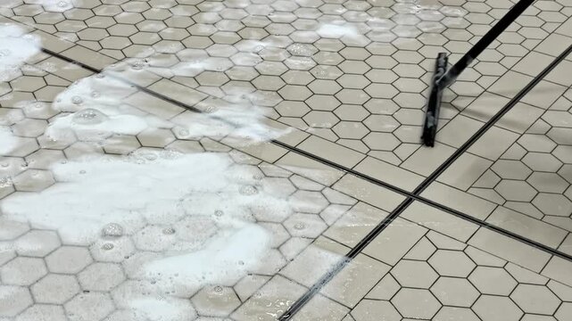 Worker cleaning tiled industrial floor with water and foam during maintenance process, squeegee