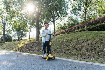 A young boy is enjoying riding a scooter in a park under bright sunlight, surrounded by greenery.