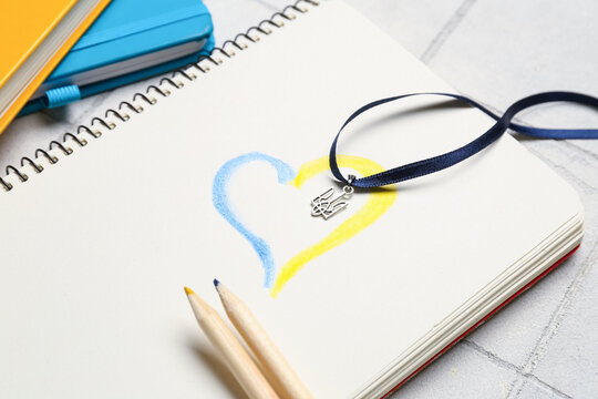 Ribbon with Ukrainian coat of arms, notebook, drawn heart and pencils on white tile background, closeup