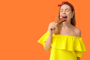 Young woman biting chocolate covered ice cream on orange background