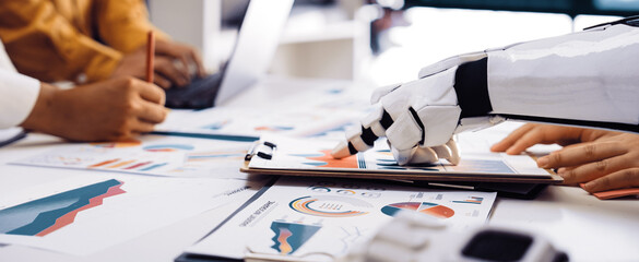 A robotic hand assists professionals in analyzing data and creating business strategies through charts and graphs in a modern office setup, showcasing technology integration.Noogenesis