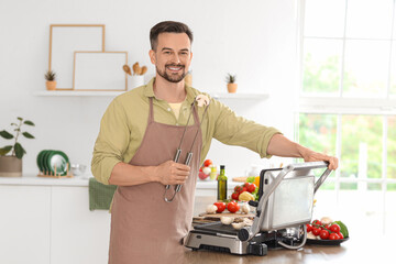 Young happy man cooking tasty fresh mushrooms on modern electric grill in kitchen