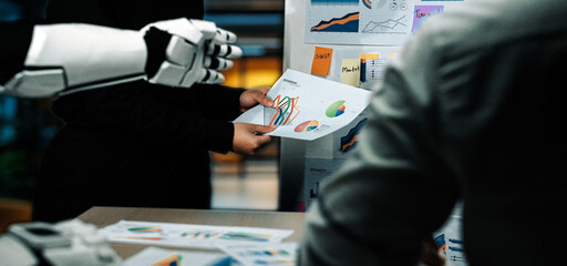 A striking image of a human and robot collaborating in a modern office setting, showcasing data analysis through charts and graphs for innovative presentation techniques.Noogenesis