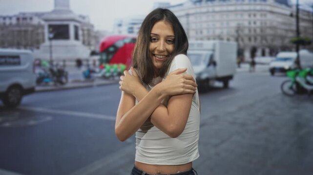 Young hispanic woman hugging bare shoulders with arms crossed on busy city street among passing vans and buses; self care contentment.