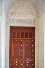 A detailed architectural photograph featuring an intricately carved wooden door framed by a beautifully decorated marble archway at the Sultan Qaboos Grand Mosque in Muscat, Oman
