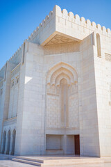 A bright architectural photograph capturing the white marble exterior of the Sultan Qaboos Grand Mosque in Muscat, Oman. The image features intricate Islamic carvings, an ornate arched doorway