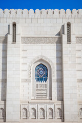 A beautifully detailed photograph showcasing the white marble fa&ccedil;ade of the Sultan Qaboos Grand Mosque in Muscat, Oman. The image highlights an ornate arched stained-glass window with geometric floral