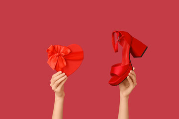 Female hands with heart-shaped gift box and heeled shoe on red background. Valentine's day...