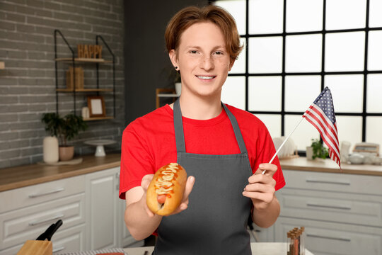 Young happy man with tasty hot dog and flag of USA in kitchen at home