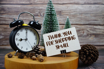 Festive holiday scene featuring a vintage alarm clock, pinecones, and miniature Christmas trees on a wooden base, with the text “Thank You For Shopping Small” encouraging support for local businesses 