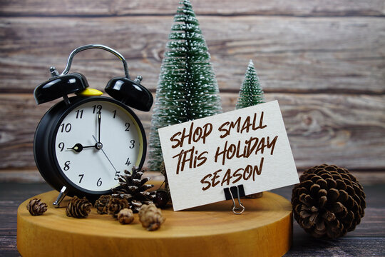 Festive holiday scene featuring a vintage alarm clock, pinecones, and miniature Christmas trees on a wooden base, with the text “Shop Small This Holiday Season” support for local businesses