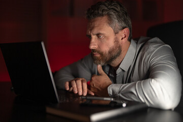 Tired businessman working late at night on a laptop in a dark office. Overworked office worker struggling with anxiety. Freelancer thinking about work. Business man concentrated on a startup project.