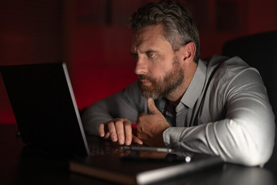 Tired businessman working late at night on a laptop in a dark office. Overworked office worker struggling with anxiety. Freelancer thinking about work. Business man concentrated on a startup project.