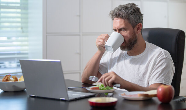 A worker eats a morning meal while checking his laptop in a cozy home office. A mature business man planning his workday. A freelancer eats a breakfast. A casual man eating lunch at home office. - Powered by Adobe