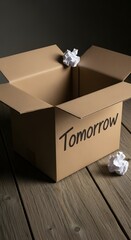 Open cardboard box with crumpled paper balls inside and the word 'Tomorrow' written on the side, placed on a wooden floor against a dark background