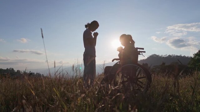 Silhouette elderly woman in wheelchair with his care helper praying to God on mountain sunset background.. Psychological medication treatment concept