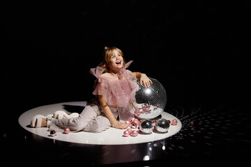Caucasian girl, about 8 years old, blonde, front view, wearing pink sheer top and sequin pants, sitting on stage floor with disco balls against dark background. Concept childhood, retro, party, show.