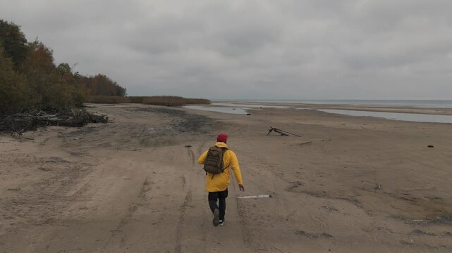 A man in a yellow jacket wearing a backpack runs along a sandy empty beach