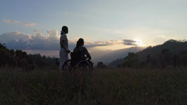 Silhouette of elderly woman in wheelchair pointing at sunset mountain to her care helper.