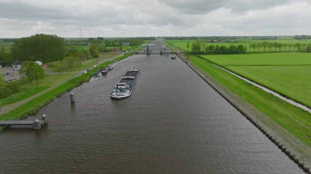 Long overview of a straight Dutch canal with barges and small craft lined up near a rural bascule bridge amid green fields.