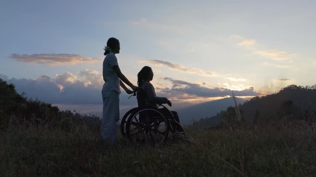 Silhouette of elderly woman in wheelchair with her care helper pointing at sunset mountain