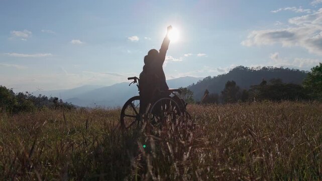 Silhouette Woman sitting on a wheelchair enjoying with raised hands on the mountain sunset time.