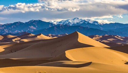 A scenic desert landscape features rolling sand dunes in the foreground, with majestic, snow-capped mountains and clouds in the background