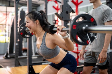 Asian woman client exercising with weight lift barbell while fitness gym trainer coach man observing