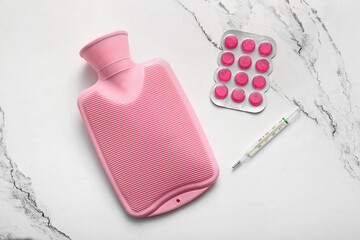 Hot water bottle with pills and thermometer on marble background