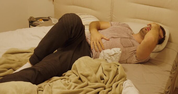A man lies curled up in bed, holding his stomach in pain, experiencing strong abdominal cramps and discomfort.