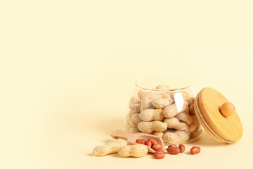 Jar with unpeeled peanuts and wooden scoop of peeled nuts on beige background