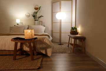 Interior of stylish bedroom with cozy bed, folding screen and glowing lamps at evening