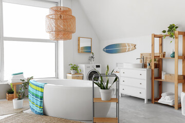 Interior of light bathroom with bathtub, washing machine, big window and surfboard
