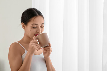 Young Asian woman drinking tea near light curtains at home