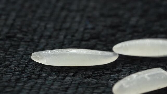 Extreme close-up 4K macro shot of raw ST25 rice grains falling in slow motion. The uncooked grains cascade onto a minimal black textured fabric, showing premium detail and movement.