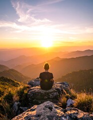 A person sits atop a rocky peak, gazing at a vibrant sunset over a series of rolling, hazy mountain ranges