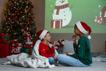 Cute children in Santa hats with popcorn watching Christmas cartoons on projector screen at home