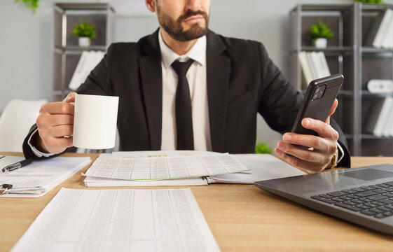 Cropped shot of serious businessman in suit holding smartphone and coffee mug while sitting at desk in office. Focused male professional reading message and working with documents during break.