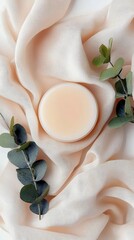 A cosmetic product, likely a balm or cream, is presented on a soft, flowing fabric with a neutral tone, accented by sprigs of eucalyptus.