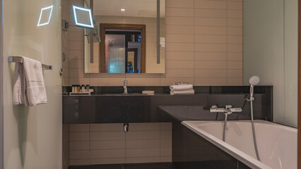 Bathroom interior. White rectangular bathtub, metal faucet, shower. The walls are tiled. Towels are on the counter in a roll, toiletries are laid out. Wall mirror.