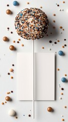 A single cake pop decorated with white frosting and multicolored sprinkles is centrally placed on a white stick, surrounded by small candies and a blank white c