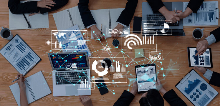 A dynamic business meeting scene showcasing professionals engaged in data visualization, interaction with technology, and strategic planning, surrounded by charts and documents. Trope - Powered by Adobe