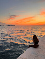 rear view of woman sitting alone on the promenade watching sunset sky over Adriatic Sea in Zadar, Croatia