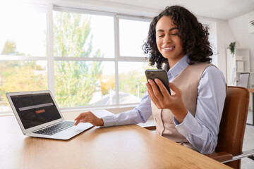 Young African-American businesswoman using mobile phone and laptop to search online at desk in...