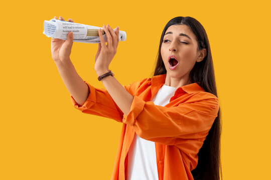 Shocked young woman looking through rolled newspaper on yellow background. Search concept - Powered by Adobe