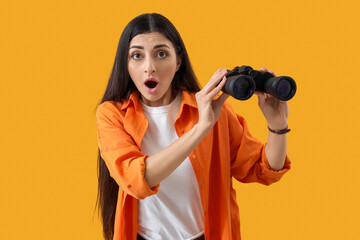 Shocked young woman with binoculars on yellow background. Search concept