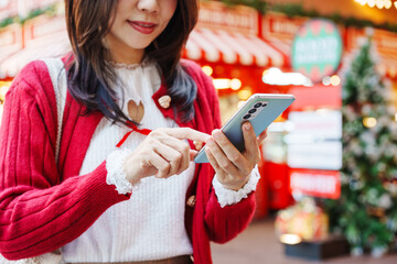 Smiling young Asian woman wearing red cardigan and using smart phone at Christmas market